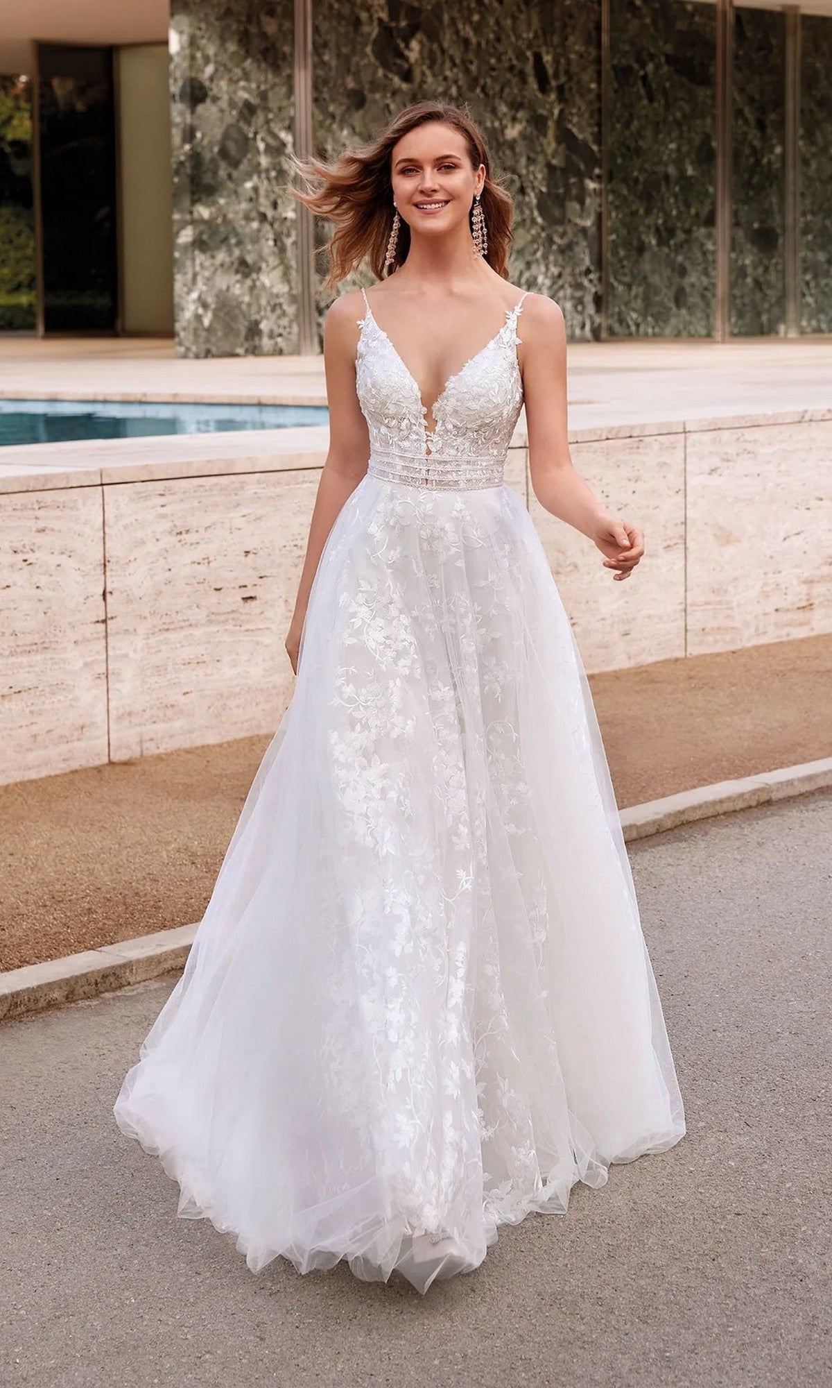 An ivory, sleeveless Long White Dress: Alyce 7036 with a deep V-neckline and floral appliqués is worn by a woman standing outside near a modern building and pool.